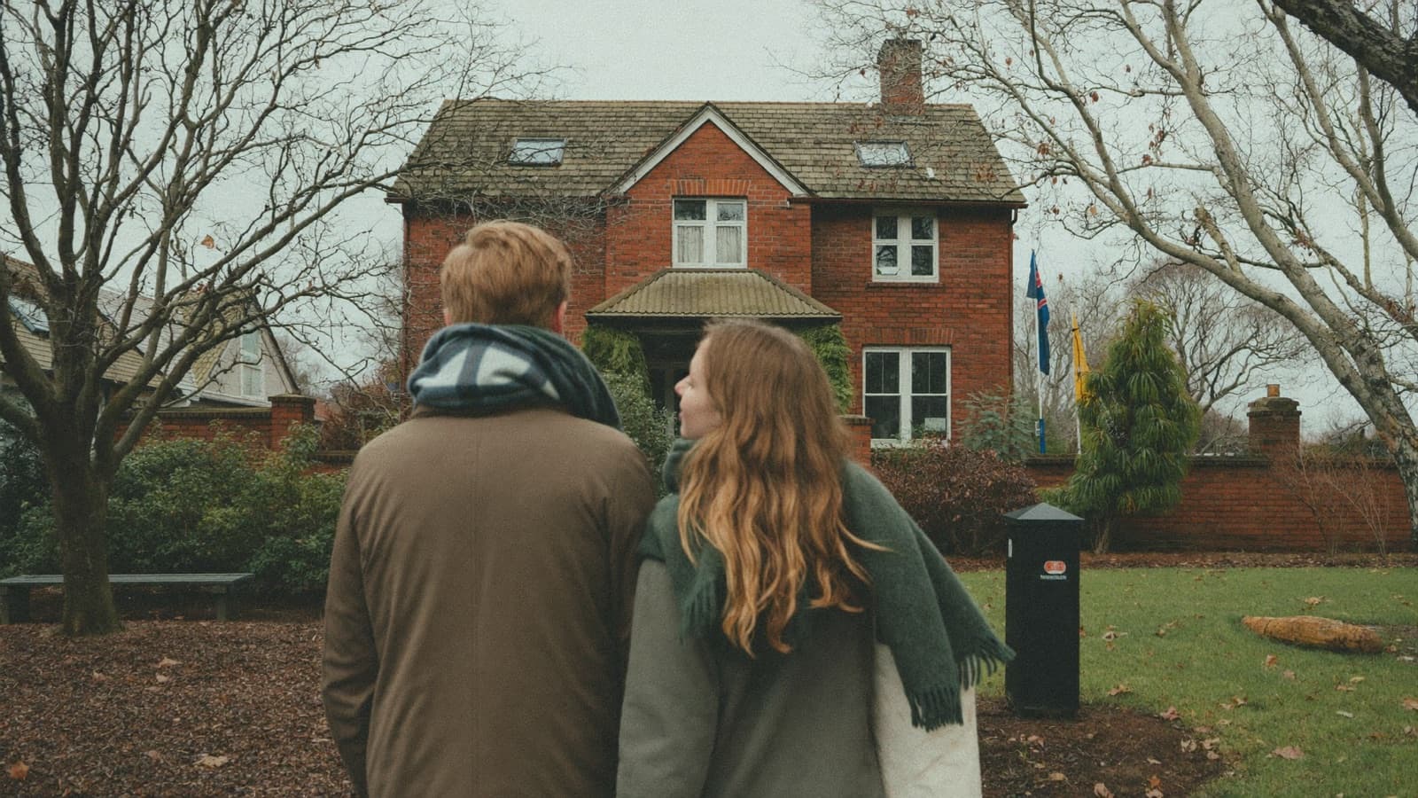A young couple in winter coats on the footpath outside a suburban brick house at an open home.