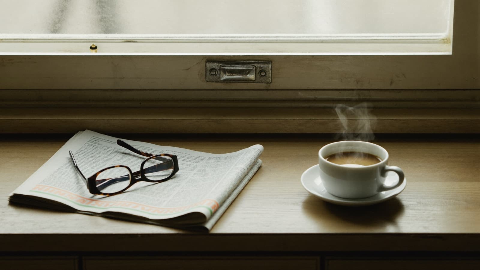 Morning still life of a folded newspaper, reading glasses and a flat white on a kitchen bench in winter light.